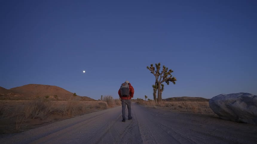 Backpacker hikes along dirt road past boulder and desert toward low mountains under clear sky with moon