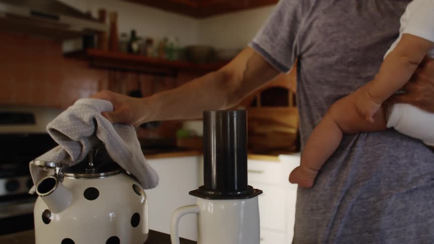 Mother uses towel to pour hot water while making coffee while holding her child