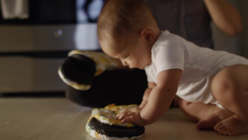 A baby plays with an oven mitt on the kitchen floor while his mother talks on her smartphone next to him