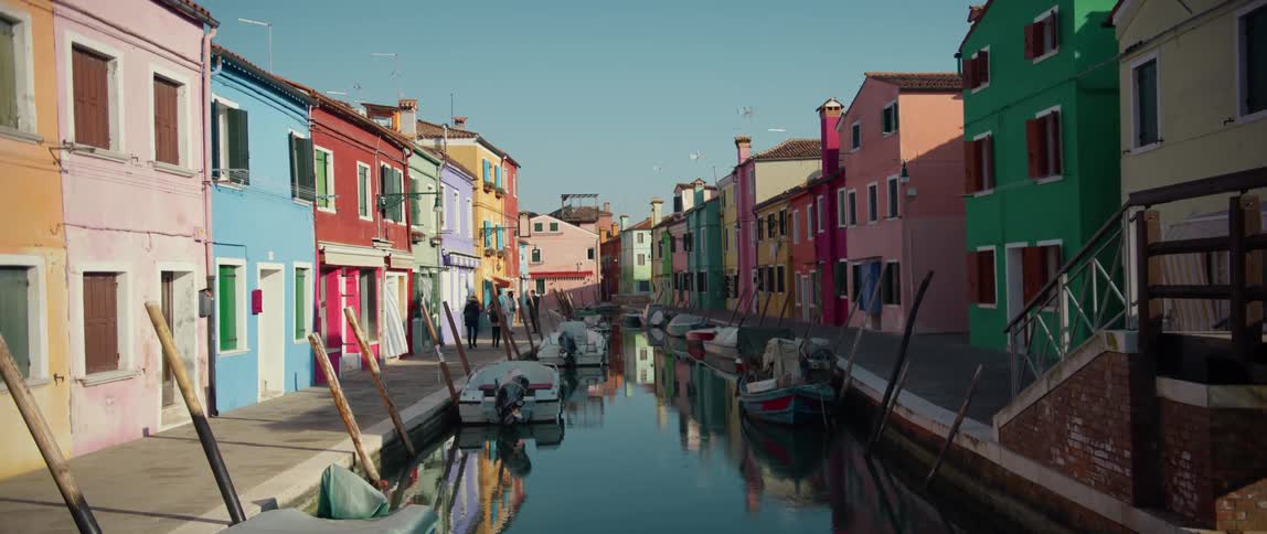 People walking along the canal past brightly colored homes, shops and boats on Burano Island, Venice, Italy