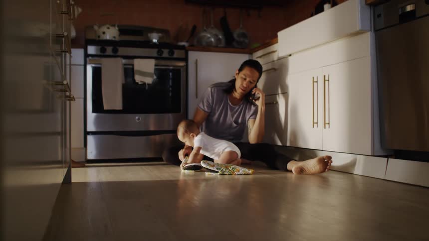 A man talks on the phone and plays with his son on the floor of his kitchen while his wife boils water