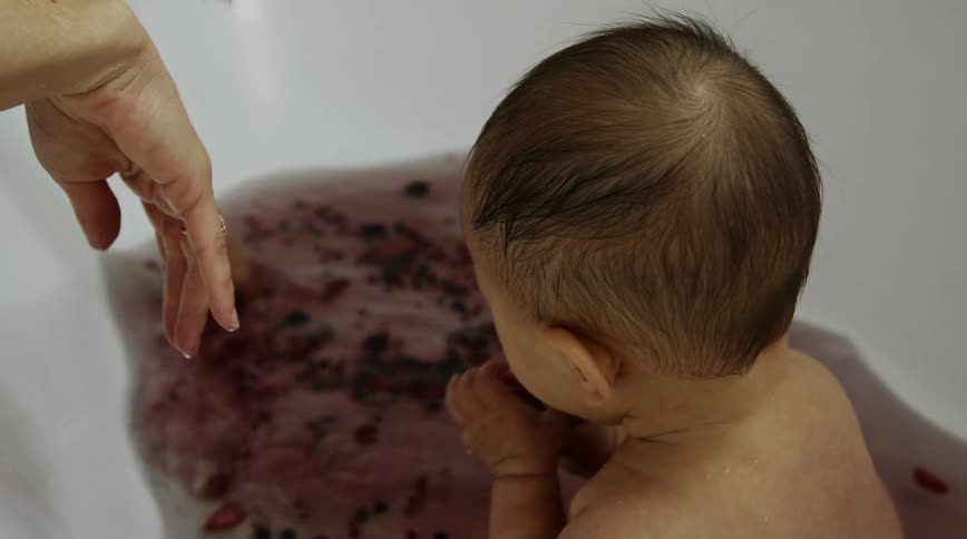 A baby sits in a tub of water with berries in as his mother stirs the water