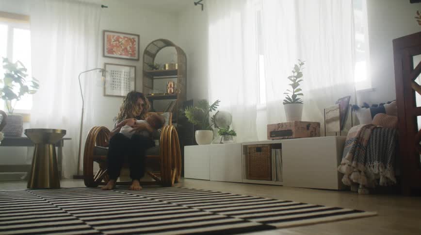 Mother with curly hair sits in armchair holding baby in living room at home