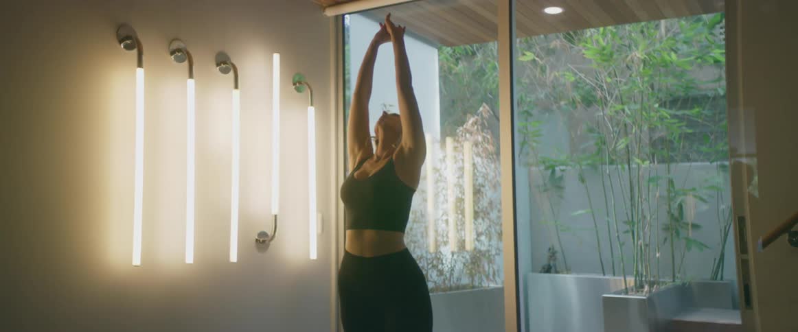 Blonde woman stands and stretches near parallel fluorescent light bars and glass doors