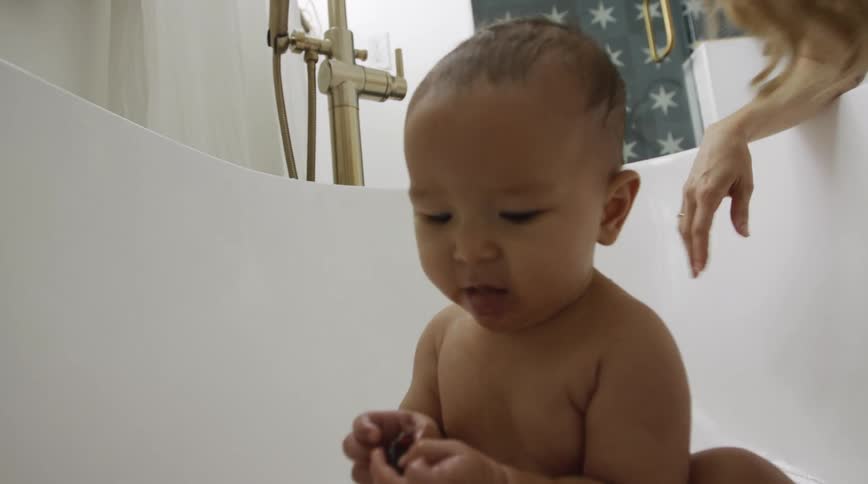 Mother watches as baby sitting in tub with fruit eats and picks up another piece