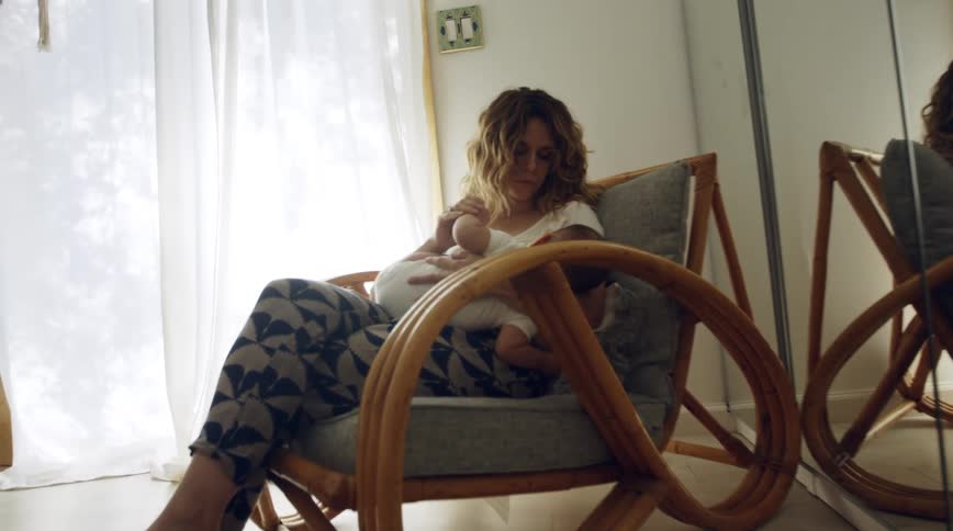 Mother with white shirt and gray pants sits in armchair and holds baby in living room at home