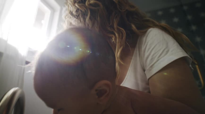 A woman laughs as she holds her baby by a sink and puts some running water on him