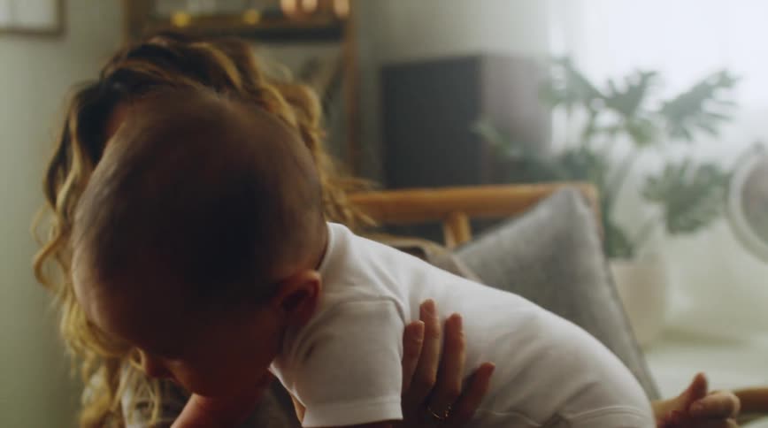 A woman at home rubs her babys shirt as she holds him and he climbs on her