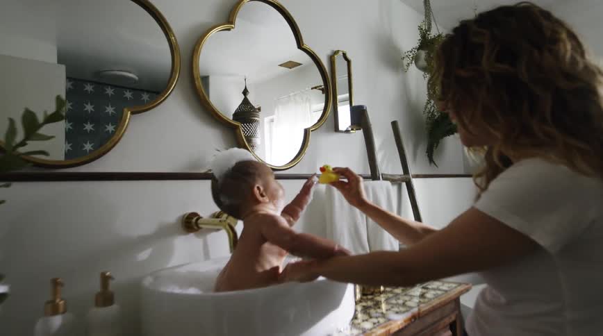 A woman uses a rubber ducky to play with a baby sitting in a baby bath tub