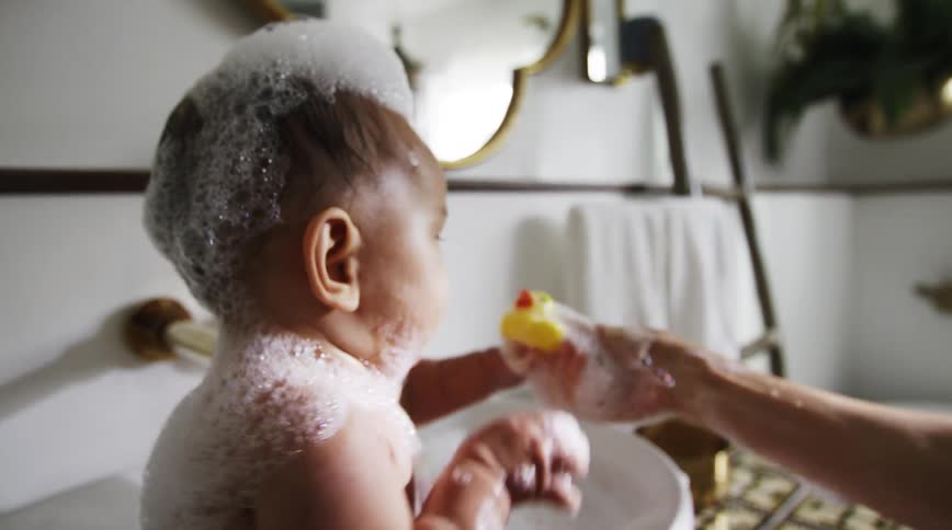 Child covered in soap foam and mother play with rubber duckie during bath time