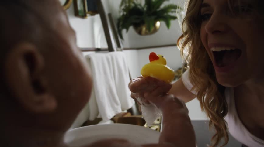 Mother hands rubber duck to child covered in soap suds