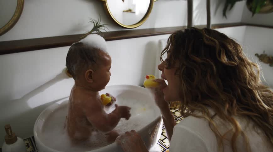 A baby sits in a sink full of water and bubbles and holds a rubber duck as his mom leans forwards and squeaks another duck at him