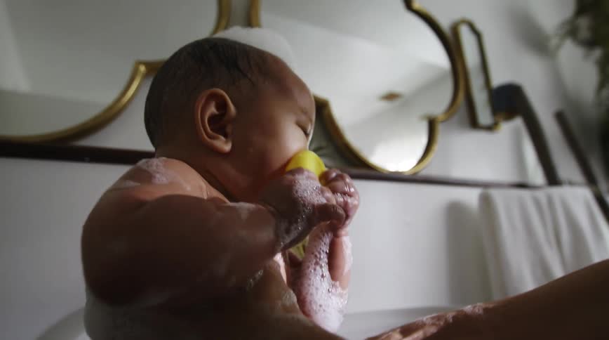A baby chews on a rubber duck as his mother steadies him with her hand while bathing him