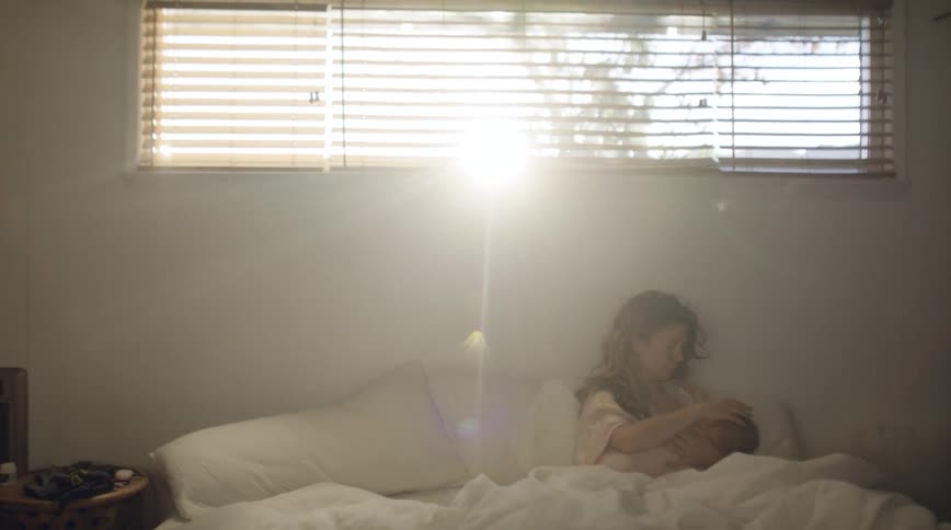 Mother with brown wavy hair sitting on bed breastfeeds baby in bedroom