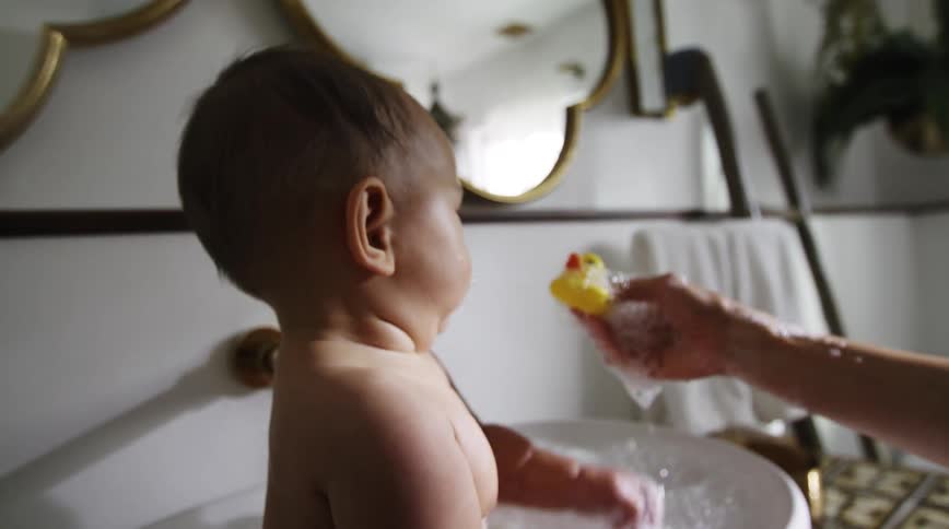 Baby pulls back looking unsure when mom bops him on the nose with a rubber duckie in the bath