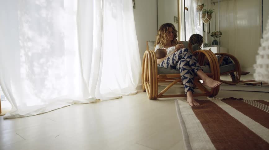 A woman sits in a chair at home with her baby in her lap and holds his hand and looks down at him