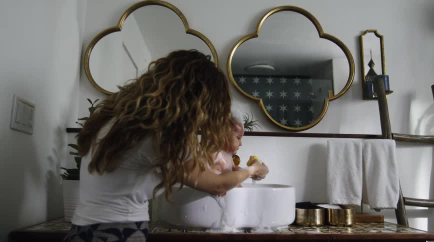 A woman squeaks a rubber duck for her baby as she bathes him in a sink