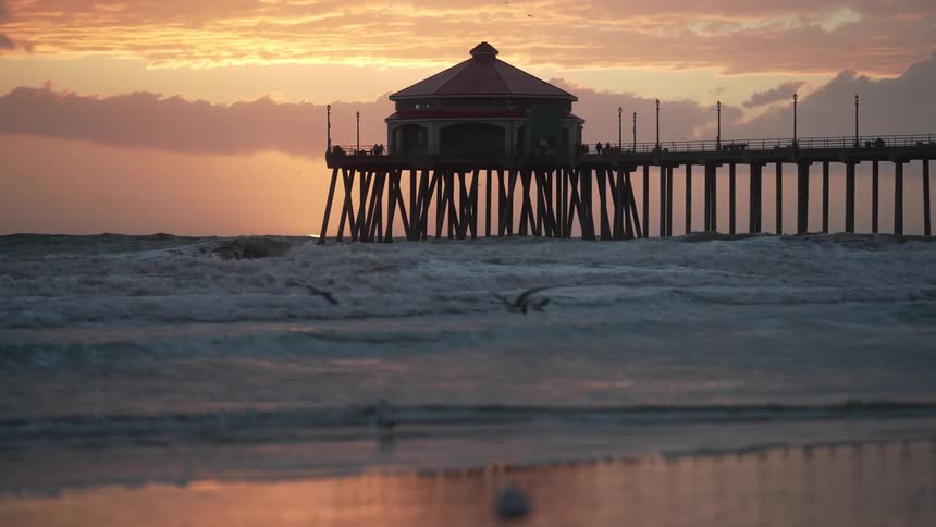 Seagulls fly over ocean waves near a pier at sunset