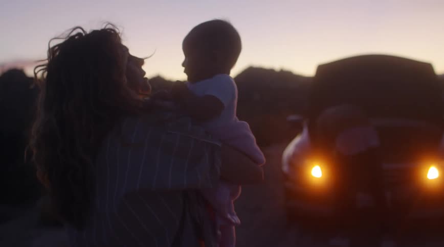 Mom carries baby to their broken down car on the side of the road where dad is bent over with the hood up