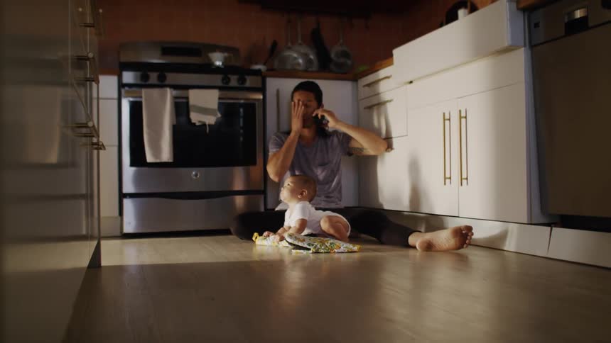 A father is talking on a cell phone while sitting on the kitchen floor with his baby