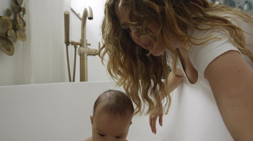 A woman reaches down in to a bathtub as she bathes her infant son