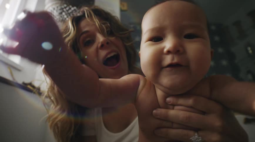 Mother with brown wavy hair and white shirt holds baby trying to reach camera in the kitchen