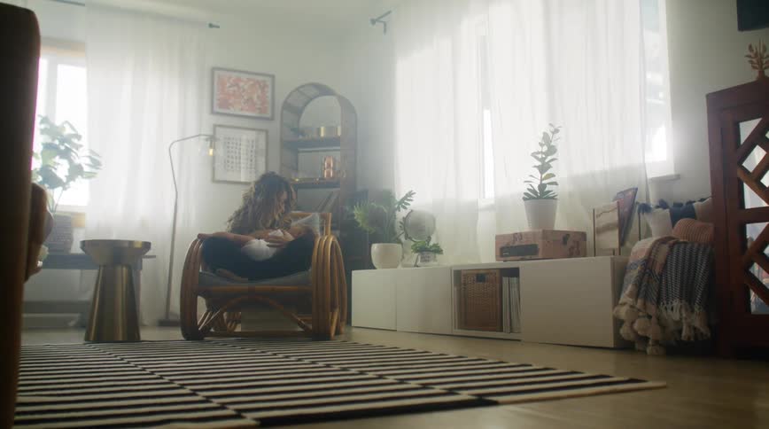 Mother with brown wavy hair black leggings sits in armchair and breastfeeds baby in living room at home