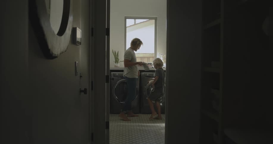 A man stands in the laundry room at home with his son and shows him how to fold something and then they both set it on the washing machine
