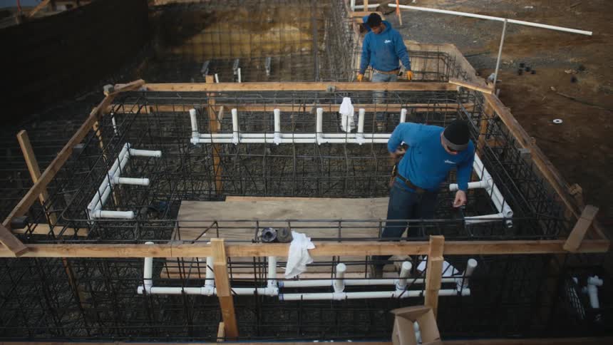 One man wearing sunglasses and a blue shirt measures with a measuring tape while another man in a blue shirt works behind him on a construction site