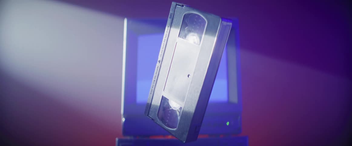A vhs tape floats and turns in a beam of light past an old computer with static on the monitor