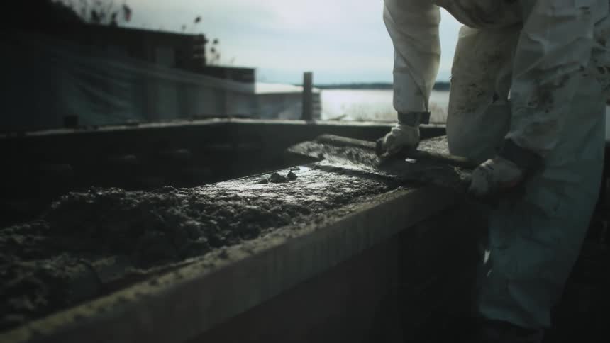 Worker in white coveralls uses a scraper to scrape wet cement on a ledge of a building