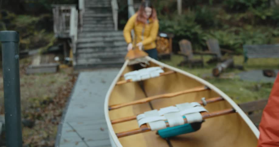 Two women wearing raincoats each hold an end of a canoe as they carry it