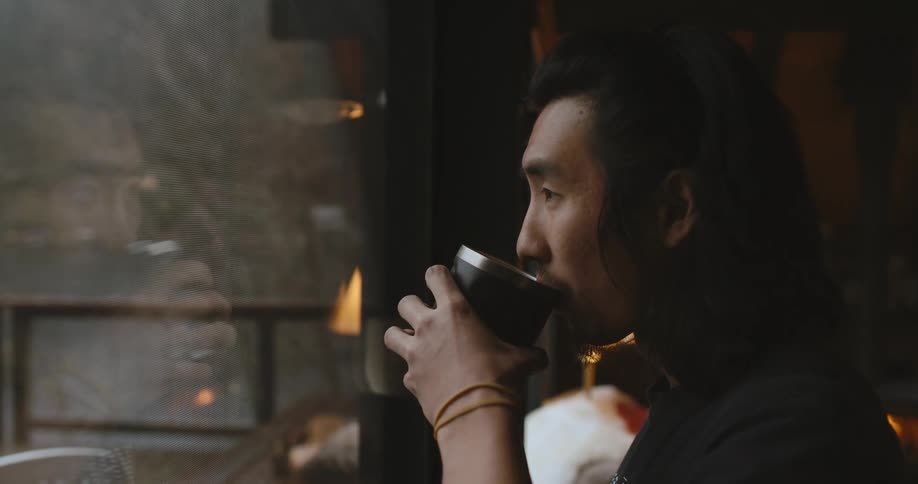 Man stands looking out window while drinking from steaming cup as fire burns in fireplace in the background