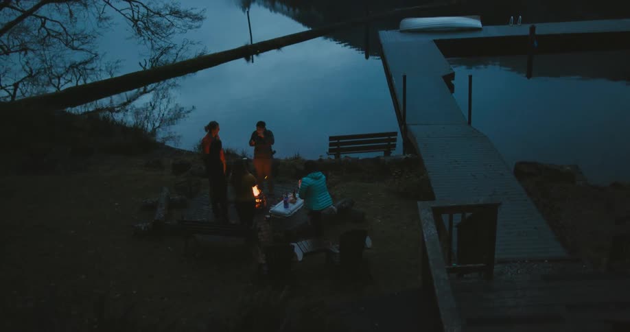 A group of women picnic around a campfire on the shore of a lake in the late evening