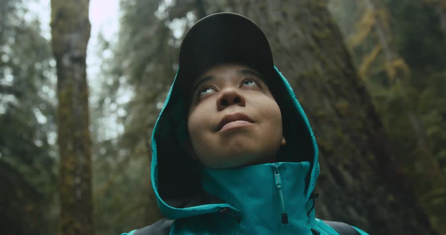 A woman wearing a raincoat with hood looking up at the trees in the rain