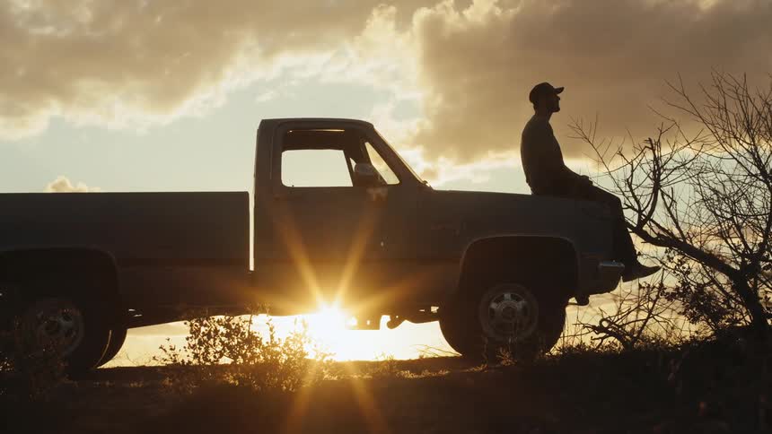 Silhouette of a man wearing a baseball cap sitting on the hood of a pickup truck as the sun shines brightly behind it