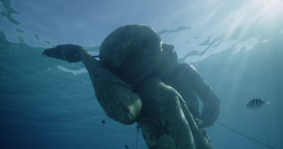 Fish swim around sculpture as sun rays shine through water from the surface