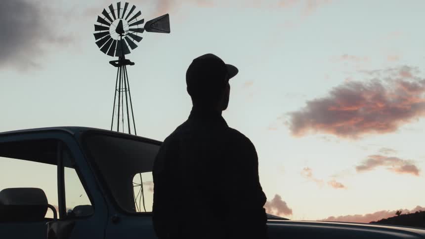 Silhouette of a man wearing a baseball cap looking at the sky and a weather vane windmill nearby