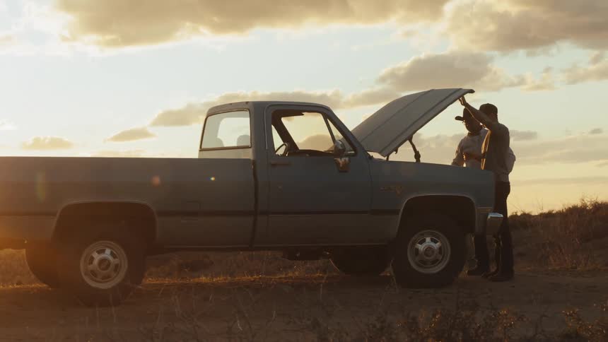 Two men open up the hood of a truck in a rural area and look inside