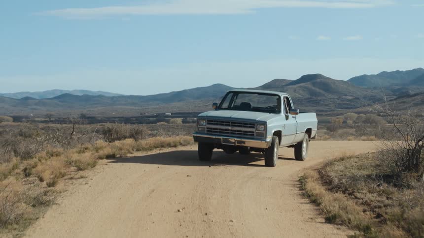 An old pickup truck is traveling on a dirt road in the desert and there are mountains in the distance