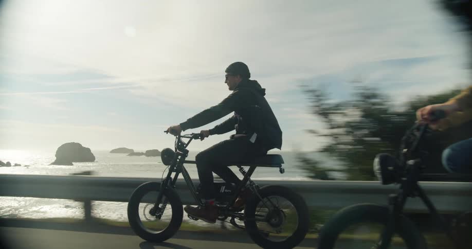 A pair of men ride motorized bicycles down a road along the coast during the day