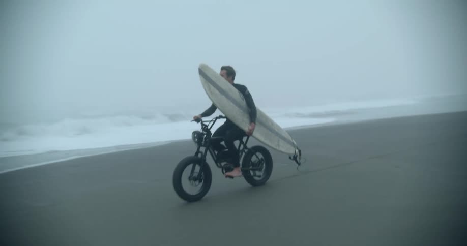 Two men carrying surf boards ride their bikes down the beach