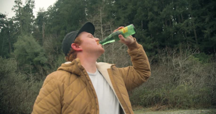 A man starts his motorized bike so the tire spins and his friend holds a bottle of water against the tire to open it and takes a drink