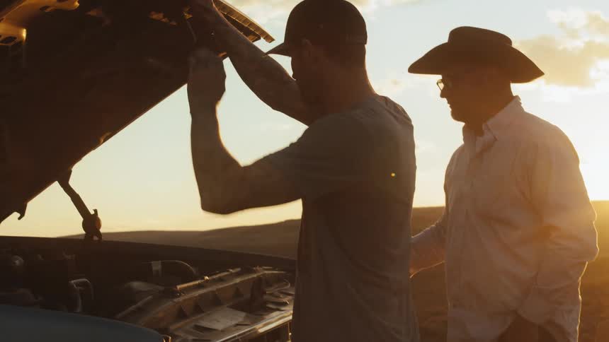 Two men in a rural area look under the hood of a truck and then one man closes it