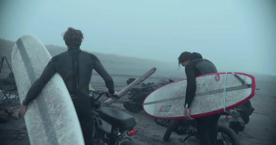 Two men holding surf boards ride up on motorcycles and greet man at camp near van on the beach