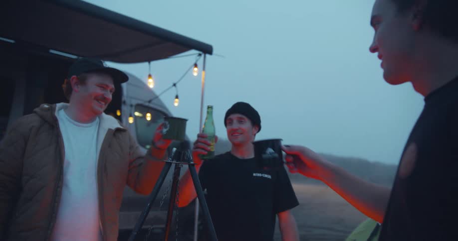 Three friends out camping click their drinks together and then one man throws his drink at another man