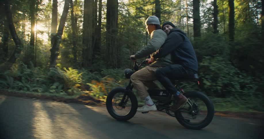 Two men with wooly har jackets and pants ride bike next to forest at sunset