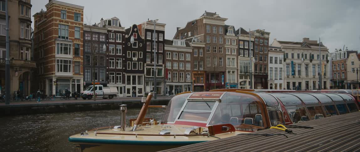 Vehicles, boats, and pedestrians travel a city street along a river canal in Amsterdam
