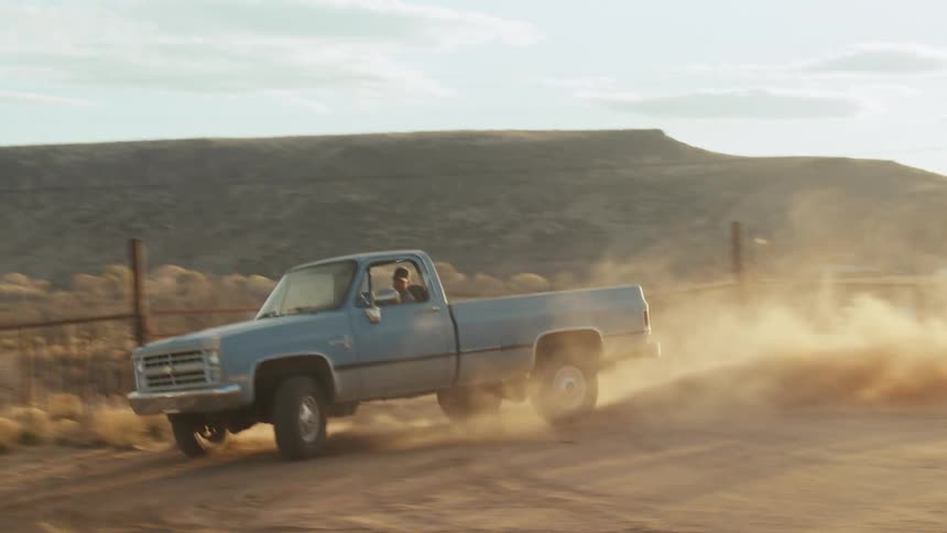 A man is driving an old pickup truck on a dirt road in the desert and kicks up a cloud of dust as he does a uturn