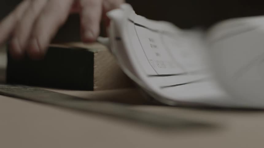 Man sorting through his drawings then using a piece of wood to hold some flat